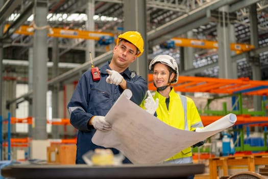 Factory workers man and woman discuss about the project using drawing paper or plan in workplace with machine and product shelves are in the background.