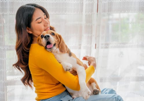 Close up young Asian girl hold and hug beagle dog and sit in front of glass door in her house and she look happy to play fun together.