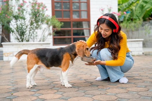 Young Asian girl with red earphone use dog bowl to give water for her pet in front of the house with day light.