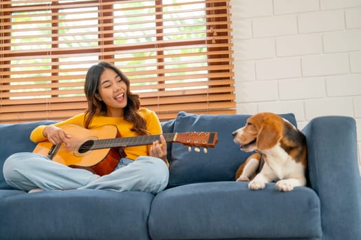 Front view of Young Asian girl sit on sofa with beagle dog to play guitar and look relax also happy in their house and dog look at the owner.