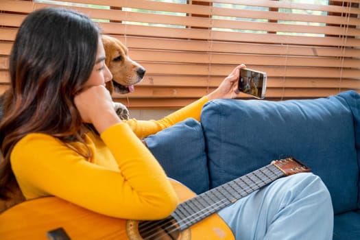 Young Asian girl enjoy to selfie with her dog during relax after play guitar on sofa in her house with day light.