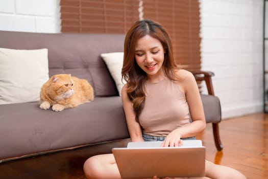 Asian pretty woman sit and use laptop to work with happiness near her orange cat that look to the owner in living room of the house.