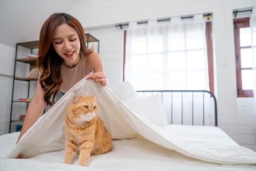 Beautiful Asian woman hold blanket over orange cat stand on bed and she look fun and happy to play with her pet in the house.