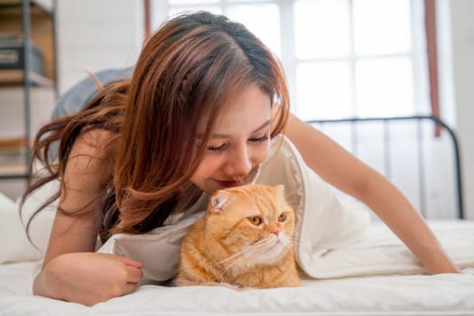 Close up beautiful Asian woman lie near her orange cat and kiss the cat that lie on bed with relax and happy emotion.