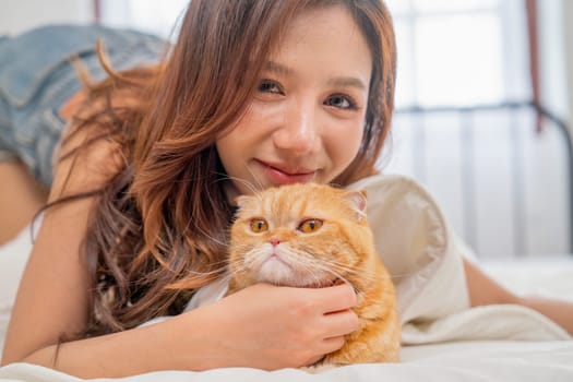 Close up beautiful Asian woman lie near her orange cat also scratch the chin of cat to make it relax and she also look at camera with smiling.