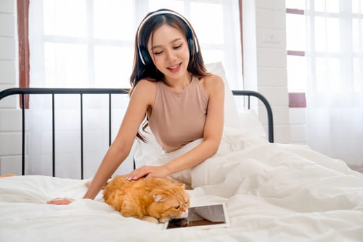 Beautiful Asian woman with headphone sit and pat orange cat that lie over tablet on bed and they look happy to stay together.