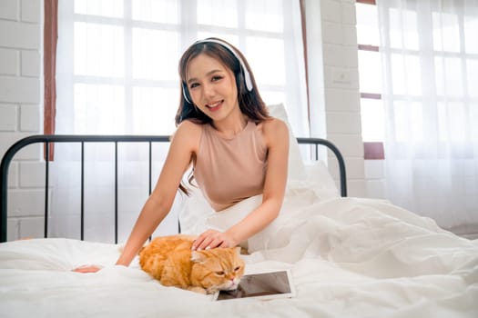 Beautiful Asian woman with headphone sit and pat orange cat that lie over tablet on bed and she also look at camera with smiling.