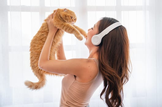 Asian girl wear headphone and hold orange cat in front of glass windows with white curtain and they look happiness together in their house.