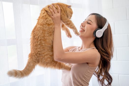 Pretty Asian girl wear earphone and hold cat in front of glass windows with white curtain and they look happiness together in their house.