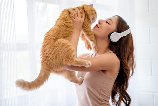 Beautiful Asian girl wear headphone and hold orange cat in front of glass windows with white curtain and they look happiness together in their house.