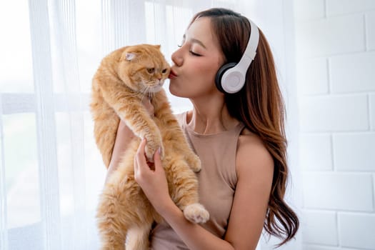 Pretty Asian girl wear earphone and hold and kiss cat in front of glass windows with white curtain and they look happiness together in their house.