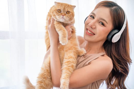 Pretty Asian girl wear earphone also look at camera hold cat in front of glass windows with white curtain and they look happiness together in their house.