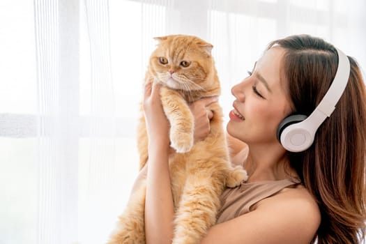 Pretty Asian girl wear earphone and hold and hug cat in front of glass windows with white curtain and they look happiness together in their house.