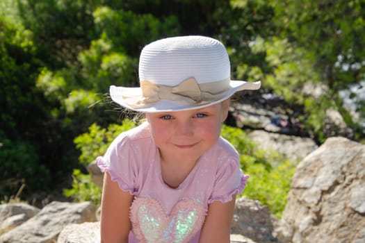 five-year-old girl in white hat smiles. She is wearing pink t-shirt, green trees in background, blue eyes, blonde hair,