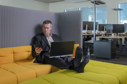 employee relaxes in IT office chillout area. He is shoeless, his feet resting on couch. office can be seen in background.