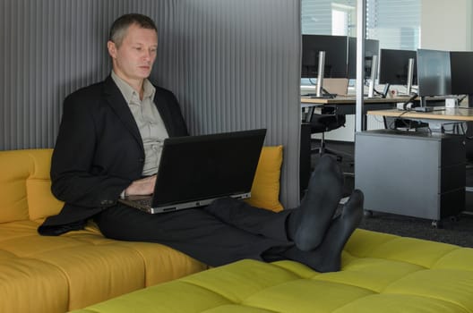 employee relaxes in IT office chillout area. He is shoeless, his feet resting on couch. office can be seen in background.