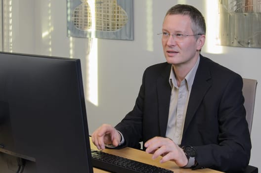 man works at computer in IT office. Middle-aged man in glasses and jacket. monitor in foreground. screen is not visible. On wall behind him glare of sun.