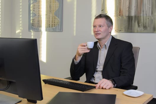 middle-aged man ina jacket holds cup of coffee in his hand. monitor is in foreground. screen is not visible. On wall behind him glare from sun.