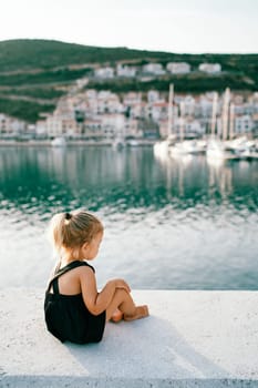 Little girl is sitting on the pier and looking at the sea. Side view. High quality photo