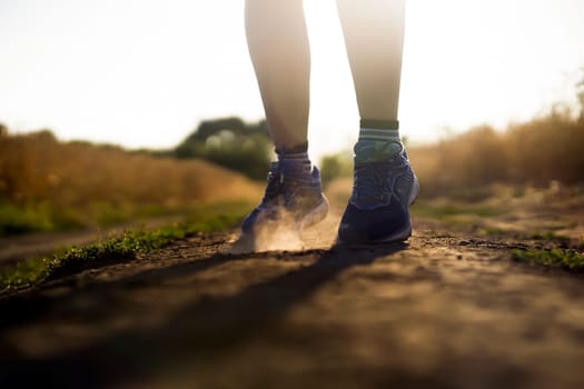 Female athletic legs in sneakers at sunset during outdoor jogging at sunset. A young girl warms up, she is engaged in trail running, view on her legs and clouds of dust close-up.