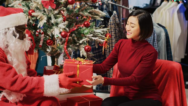 Customer in fashion store talking with actor dressed as Santa Claus, sitting next to Christmas tree during festive promotional event. Client receiving present from worker during holiday season