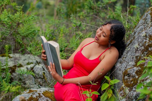 latina girl in red dress leaning against a tree surrounded by nature with an open book in her hands. High quality photo