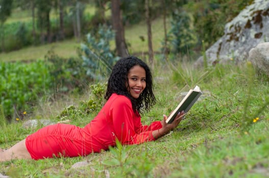 beautiful latin american woman with black hair lying face down on the grass of a mountain in a red dress with an open paper book and a beautiful smile. book day. High quality photo