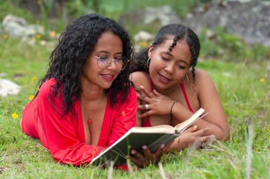 two latina friends lying face down together reading a book attentively the girl holding the book has a pair of glasses. book day. High quality photo
