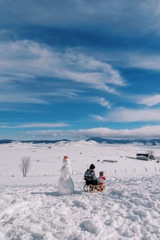 Mom with a small child sit on a sled near a snowman and look at the snow plain. Back view. High quality photo