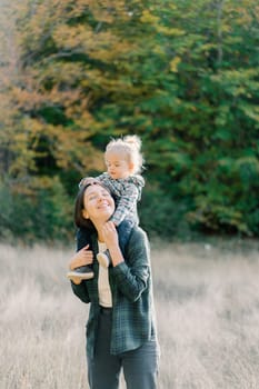 Little girl touching her mother head while sitting on her shoulders in the park. High quality photo