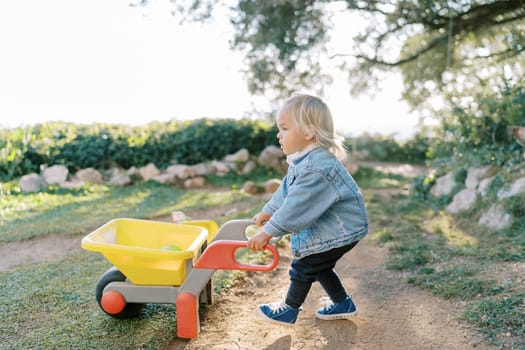 Little girl with a toy wheelbarrow walks along a dirt path in the park. High quality photo