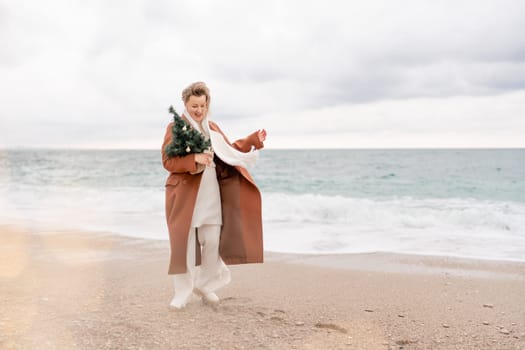 Blond woman Christmas sea. Christmas portrait of a happy woman walking along the beach and holding a Christmas tree in her hands. She is wearing a brown coat and a white suit