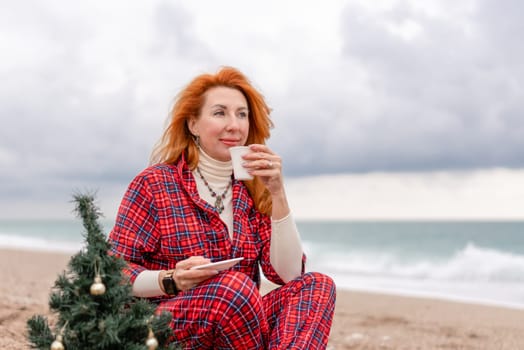 Sea Lady in plaid shirt with a mug in her hands enjoys beach with Christmas tree. Coastal area. Christmas, New Year holidays concep.