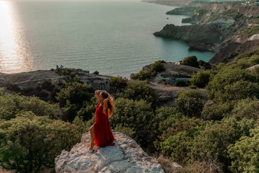 Woman sunset sea red dress, side view a happy beautiful sensual woman in a red long dress posing on a rock high above the sea on sunset