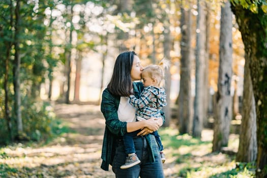 Mom hugs and kisses on the forehead a little girl holding her in her arms in the autumn park. High quality photo