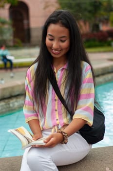 young girl from latin america sitting at the edge of a public fountain filled with water reading a book while smiling and enjoying her reading. book day. High quality photo