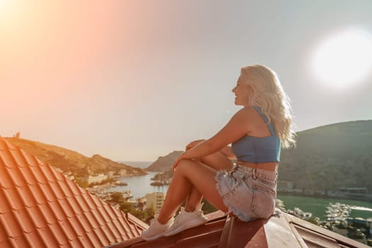 Woman sits on rooftop, enjoys town view and sea mountains. Peaceful rooftop relaxation. Below her, there is a town with several boats visible in the water. Rooftop vantage point