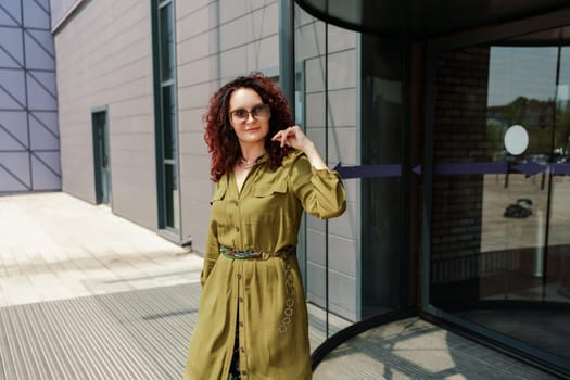 Portrait of a woman standing near a supermarket building. Caucasian model with long brunette hair, wears sunglasses and a khaki dress