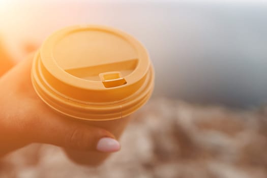 Hand holding Yellow cup with lid, coffee against a backdrop of a blue sky and sea. Illustrating cup and beverage.