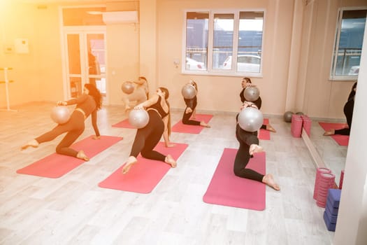 A group of six athletic women doing pilates or yoga on pink mats in front of a window in a beige loft studio interior. Teamwork, good mood and healthy lifestyle concept