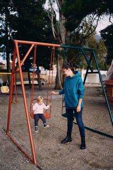 Mom stands next to the swing on the playground, with a little girl sitting on it. High quality photo
