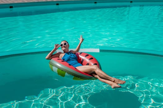Happy woman in a swimsuit and sunglasses floating on an inflatable ring in the form of a watermelon, in the pool during summer holidays and vacations. Summer concept