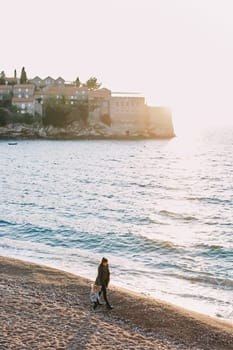 Mom and a little girl are walking along the seashore with the island of Sveti Stefan in the background. Montenegro. High quality photo