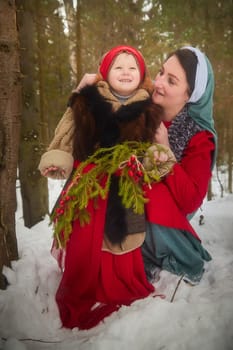 Family with mother and little daughter in stylized medieval peasant clothing in winter forest. The woman and child pose for fairy tale photoshoot in nature on a cold day. Concept of love, friendship