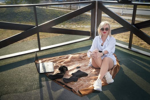 Fashionable beautiful blonde girl in a white shirt and a short skirt is resting in a gazebo with a laptop on sunny autumn, spring day. Lazy and relaxing businesswoman, manager, student in nature