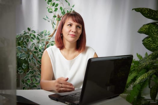 Serious girl with laptop and notebook is working at a table in cozy room with flowers. A middle-aged business woman. Psychologist in the office