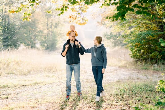 Mom touches the hand of dad, carrying a little boy on his shoulders, walking in the autumn park. High quality photo