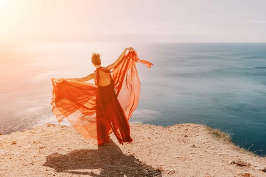 Side view a Young beautiful sensual woman in a red long dress posing on a rock high above the sea during sunrise. Girl on the nature on blue sky background. Fashion photo.