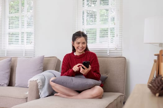 Happy young Asian woman relaxing on a soft sofa. At home, send messages via smartphone. Smiling young woman using a mobile phone online message chat Buy things online from home.