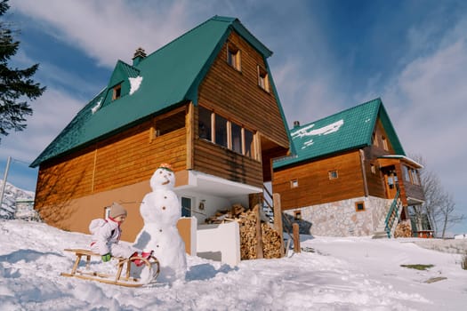 Little girl slides down a hill on a wooden sled next to a snowman near a wooden cottage. Side view. High quality photo